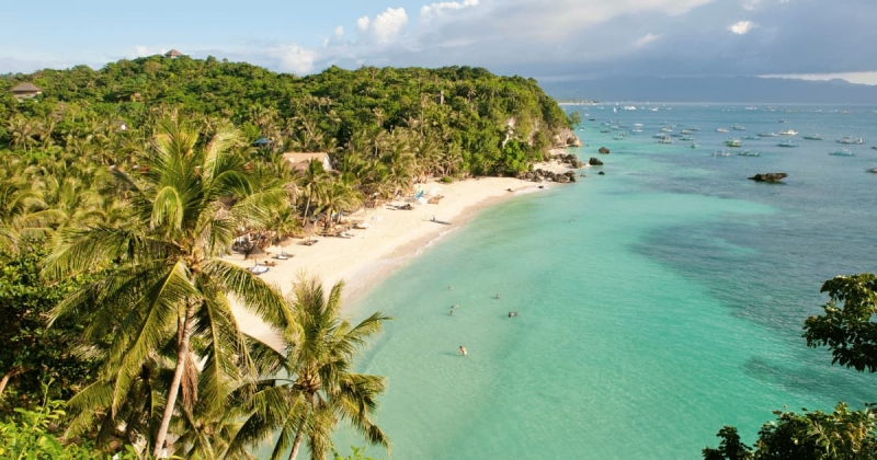A relaxing view of Boracay’s White Beach in the Philippines, featuring golden sand, clear turquoise water, swaying palm trees, and visitors enjoying a sunny tropical day