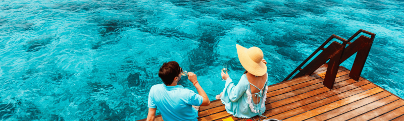 Romantic couple enjoying drinks on an overwater deck at a tropical island resort, overlooking the crystal-clear blue ocean in the Maldives.