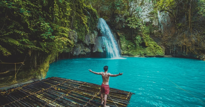 A man standing on a bamboo raft admiring the turquoise waters and cascading Kawasan Falls in Badian, Cebu, Philippines, surrounded by lush tropical jungle
