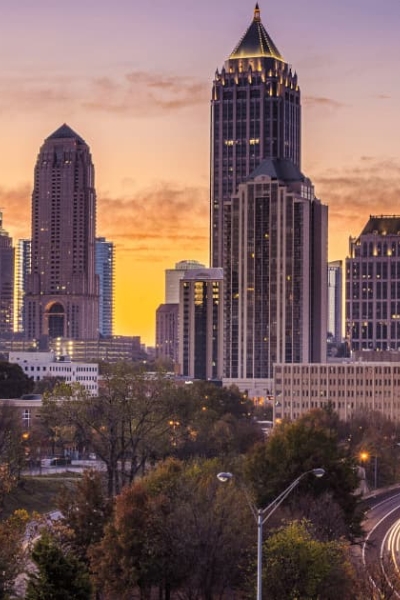 Atlanta skyline at sunset with tall skyscrapers and a curving highway lit by car lights in the foreground.