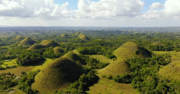 A wide aerial view of the Chocolate Hills in Bohol, Philippines, featuring hundreds of perfectly shaped green mounds under a bright sky