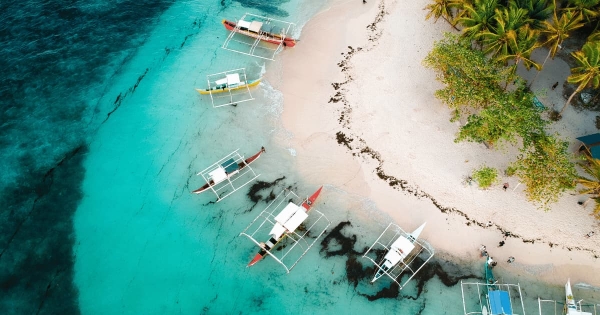 Aerial view of colorful boats floating in turquoise waters around the sandy shores of Guyam Island in Siargao, Philippines