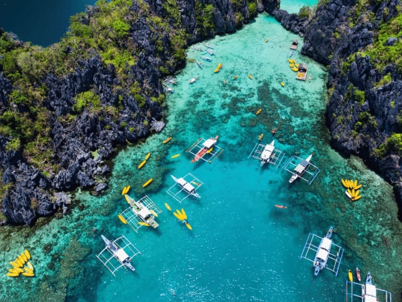 A stunning aerial view of El Nido’s turquoise lagoon in Palawan, Philippines, surrounded by dramatic limestone cliffs and traditional outrigger boats floating on crystal-clear water