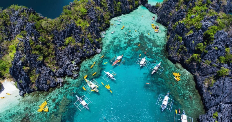 A stunning aerial view of El Nido’s turquoise lagoon in Palawan, Philippines, surrounded by dramatic limestone cliffs and traditional outrigger boats floating on crystal-clear water