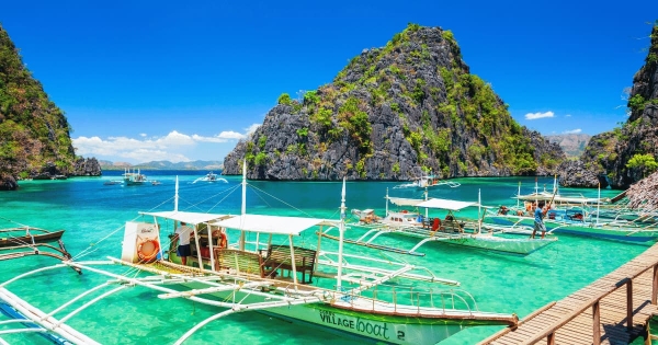 Traditional Filipino outrigger boats docked at the emerald-green waters of El Nido, Palawan, Philippines, with towering limestone cliffs and a bright blue sky in the background