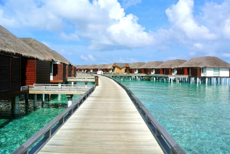 Wooden walkway leading to rows of overwater villas above clear turquoise water under a bright blue sky.