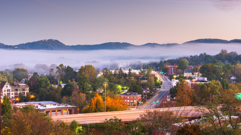 Scenic view of Asheville, North Carolina, with fog rolling over the Blue Ridge Mountains and fall foliage surrounding the city streets.