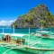 Traditional Filipino outrigger boats docked at the emerald-green waters of El Nido, Palawan, Philippines, with towering limestone cliffs and a bright blue sky in the background