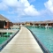 Wooden walkway leading to rows of overwater villas above clear turquoise water under a bright blue sky.
