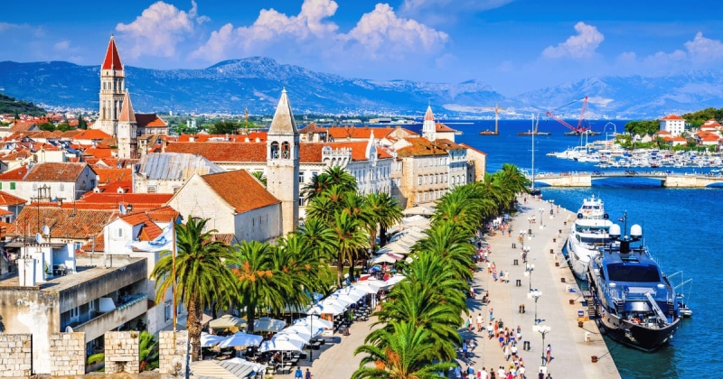 The lively promenade of Trogir, Croatia, is lined with palm trees and historic buildings, featuring the cathedral bell tower and yachts docked on the shimmering Adriatic Sea