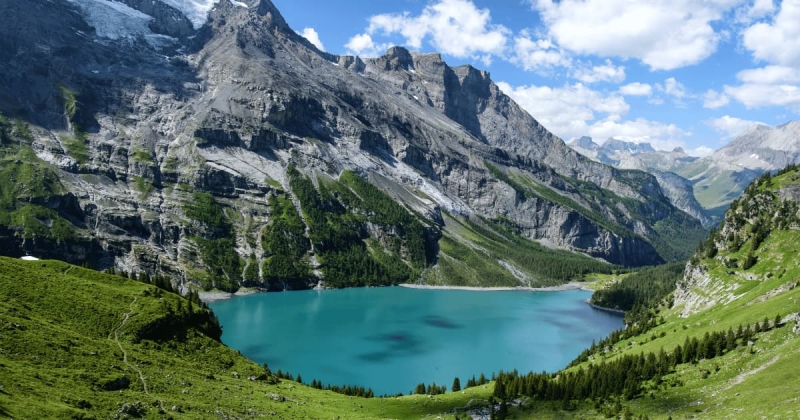 The vibrant turquoise waters of Oeschinensee lake in Switzerland are cradled by towering, rocky Alpine mountains and surrounded by bright green meadows and pine forests