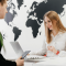 Travel agent assisting a client at an office desk with a laptop, smiling while discussing travel plans. A large world map decorates the wall in the background