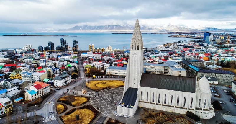 An aerial view of the Hallgrímskirkja church dominating the colorful cityscape of Reykjavík, Iceland, with the harbor and snowy mountains visible in the distance