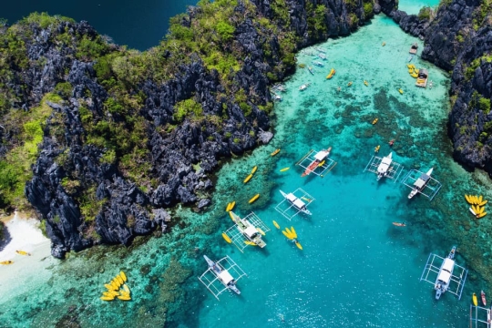 A stunning aerial view of El Nido’s turquoise lagoon in Palawan, Philippines, surrounded by dramatic limestone cliffs and traditional outrigger boats floating on crystal-clear water
