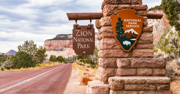 Entrance sign for Zion National Park in Utah, USA, with National Park Service emblem and scenic red rock cliffs in the background.