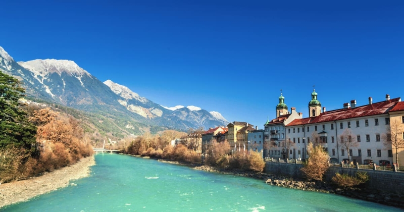 The vibrant turquoise Inn River flows past historic buildings in Innsbruck, Austria, with the steep, snow-dusted peaks of the Alpine mountains rising dramatically in the background under a bright blue sky