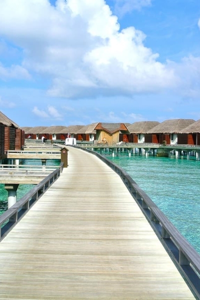 Wooden walkway leading to rows of overwater villas above clear turquoise water under a bright blue sky.