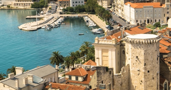 An elevated view over a boat harbor in Split, Croatia, showing the bright turquoise water, palm trees, and the ancient stone walls of Diocletian's Palace in the foreground