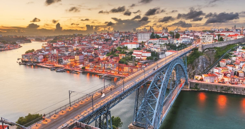 The city of Porto, Portugal, glows in the golden light of sunset, with the Dom Luís I Bridge spanning the Douro River and the colorful Ribeira district lining the water's edge