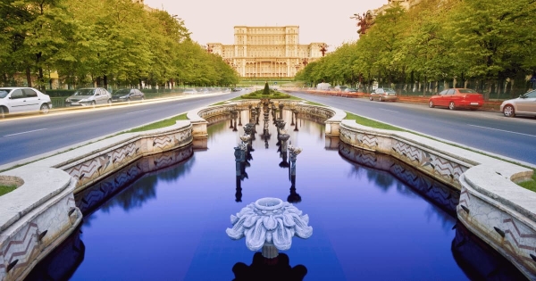 The majestic Palace of the Parliament in Bucharest, Romania, stands at the end of a grand boulevard, reflected in a central water fountain, lined by lush green trees and blurring city traffic