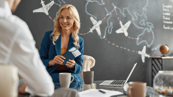 Travel agent holding flight tickets while meeting a client in an office with a world map and airplanes drawn on a chalkboard wall.