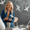 Travel agent holding flight tickets while meeting a client in an office with a world map and airplanes drawn on a chalkboard wall.