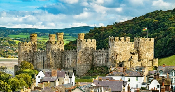 The medieval stone walls and eight round towers of Conwy Castle in Wales stand guard over the town, backed by lush green hills under a bright sky, flying the Welsh flag
