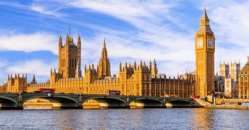 A sunny view of the Palace of Westminster and the Elizabeth Tower (Big Ben) along the River Thames in London, England, with a red double decker bus crossing the bridge