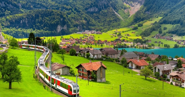A red and white passenger train winds through the lush green hillside above the village of Lungern, Switzerland, with the turquoise Lake Lungern visible in the background