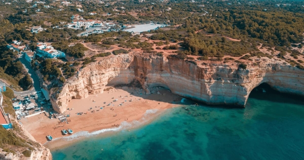 An aerial view of a secluded golden sand beach in the Algarve, Portugal, showing turquoise water meeting dramatic, towering limestone cliffs and sea caves