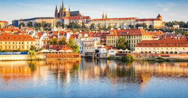 The majestic Prague Castle complex sits atop a hill, overlooking the traditional red roof buildings along the Vltava River in Prague, Czechia, perfectly reflected in the water
